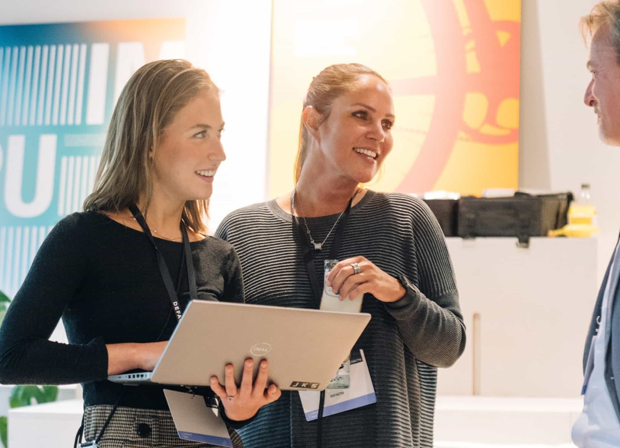 Zwei Mitarbeiterinnen von DEFACTO auf einem Event im Gespräch, mit Laptop in der Hand.